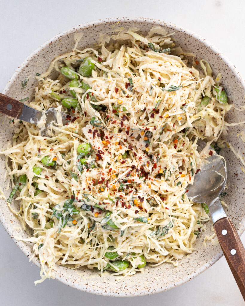 finished cabbage salad in a mixing bowl with two spoons for tossing, with crushed red pepper flakes sprinkled on top